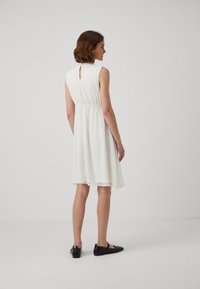 Robe blanche sans manches avec un détail de dos en trou de serrure, taille élastique et tissu texturé. Portée avec des chaussures noires, tombant au-dessus du genou.
