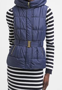 Marinblå pufferväst med hög krage, bältad midja och två blixtlåsfickor; har quiltad textur och detaljer i guld.