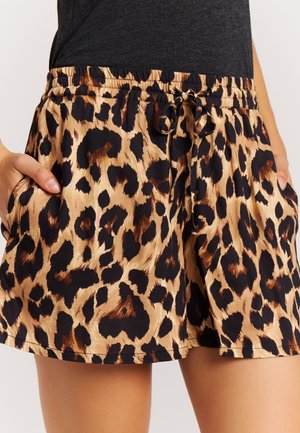 Leopardprint korte broek met elastische tailleband en trekkoord, gedragen met handen in de zakken en gecombineerd met een donker shirt.
