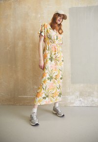 Virágos maxiruha rózsaszín, narancssárga és zöld árnyalatokban, behúzott derékkal és rövid ujjal, ezüst sportcipőkkel párosítva.