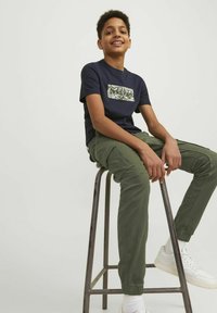 T-shirt bleu marine avec un carré de logo à motif vert, associé à un pantalon cargo vert et des baskets blanches. Un homme assis sur un tabouret en métal.