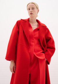 Cappotto rosso in lana con ampi rever, vestibilità over size e tasche, indossato sopra una blusa in raso rosso e pantaloni coordinati.