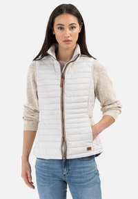 Leichtgewichtige gesteppte Weste in hellem Creme mit hohem Kragen, Reißverschluss und kontrastierendem braunen Reißverschluss, kombiniert mit einem beigen Strickpullover darunter.