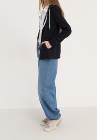 Femme debout de profil portant un sweat à capuche noir zippé, un t-shirt blanc, un jean bleu ample et des baskets beiges avec des lacets violets sur un fond uni.