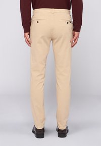 Beige Chinos mit schmaler Passform, zwei Gesäßtaschen und einer glatten Oberfläche. Getragen mit dunklen Schuhen, mit geradem Beinschnitt.