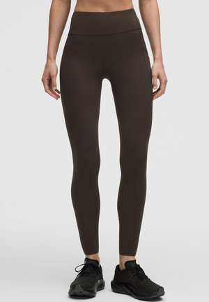 Donna che indossa leggings marroni scuro a vita alta e scarpe sportive nere, in piedi davanti a uno sfondo semplice.
