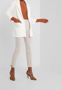 Weiße Blazer, rostfarbenes Strickoberteil, beige Hose und gestreifte High-Heels. Der Blazer hat Taschen und das Oberteil hat eine gerippte Textur.