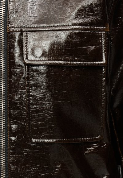 Κοντινό πλάνο τσέπης γυαλιστερού σκούρου καφέ δερμάτινου μπουφάν με πτερύγιο και ορατό ράψιμο δίπλα σε μεταλλικό φερμουάρ.