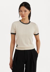 Krémový krátky pletený top s čiernym lemovaním na golieri a rukávoch, so subtilným vyšívaným logom v dolnom ľavom rohu.