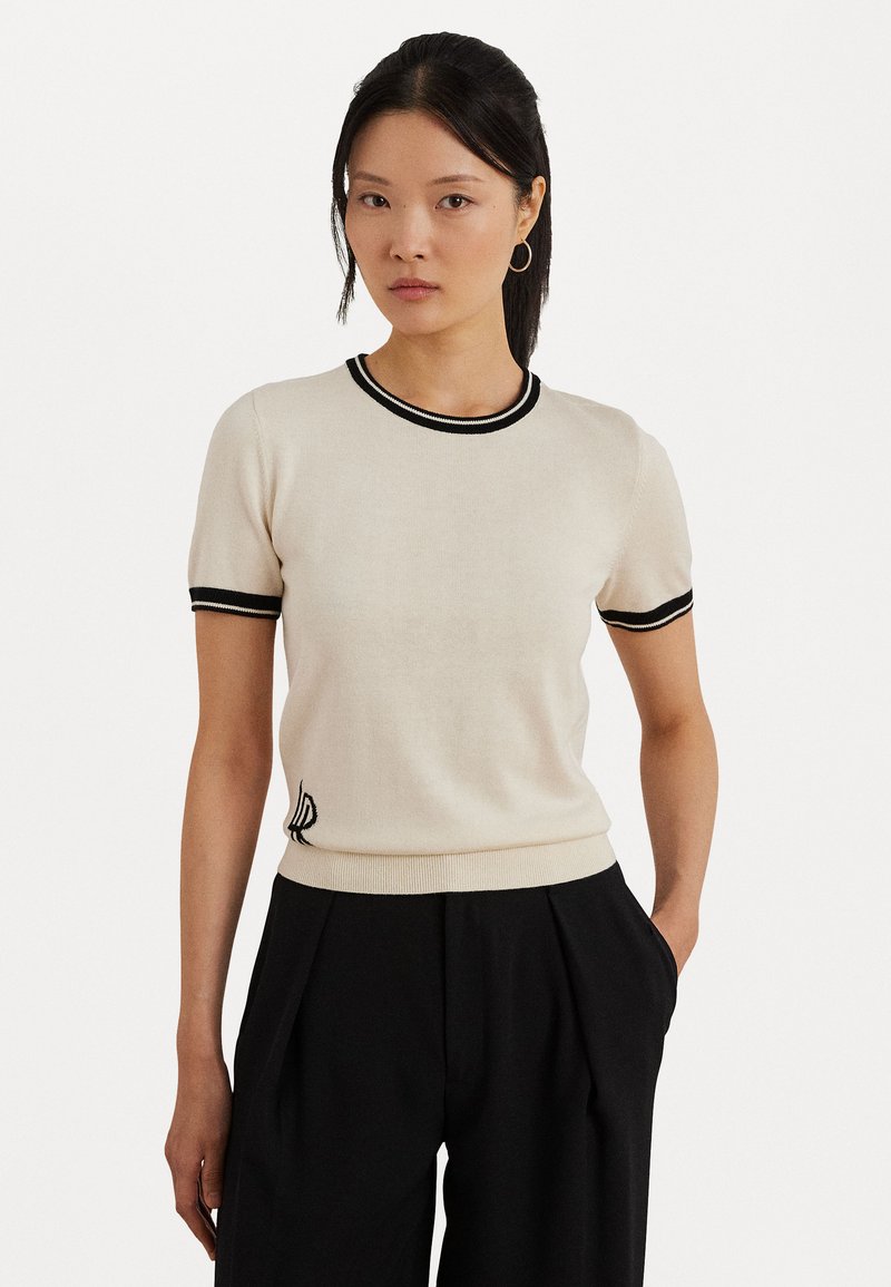 Krémový krátky pletený top s čiernym lemovaním na golieri a rukávoch, so subtilným vyšívaným logom v dolnom ľavom rohu.