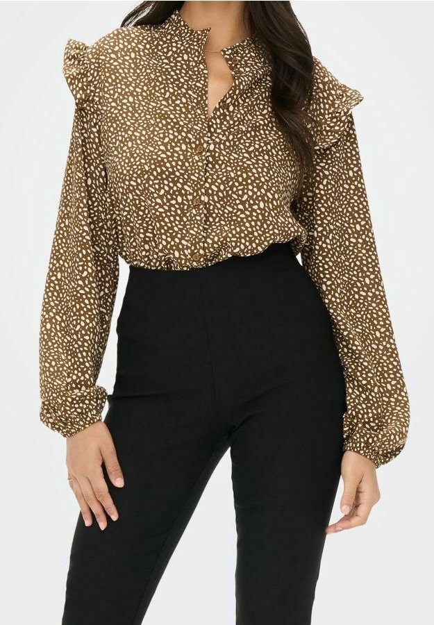 Braunbluse mit weiß gepunkteten Mustern und rüschigem Kragen. Lange Ärmel mit elastischen Bündchen. Kombiniert mit schwarzen Hosen mit hoher Taille.