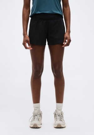 Osoba ubrana w czarne szorty sportowe, turkusowy bezrękawnik, białe skarpety za kostkę i beżowe buty do biegania stoi na jednolitym tle.