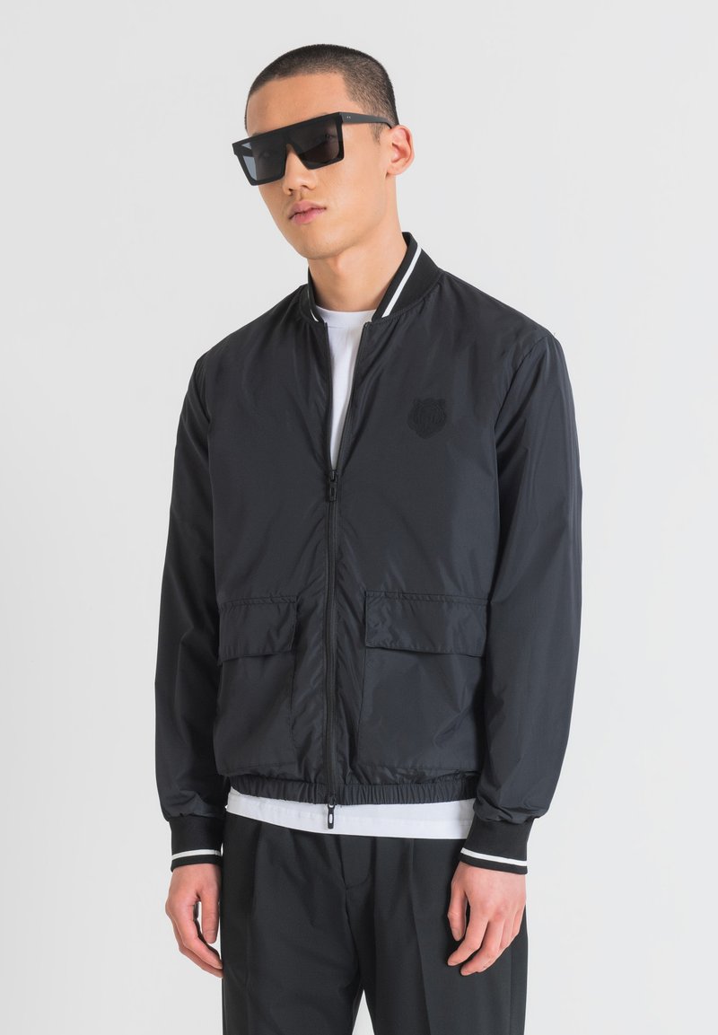 Giubbotto bomber nero con chiusura a zip, polsini a coste, due tasche anteriori e un logo discreto. Indossato sopra una camicia bianca, abbinato a pantaloni neri.