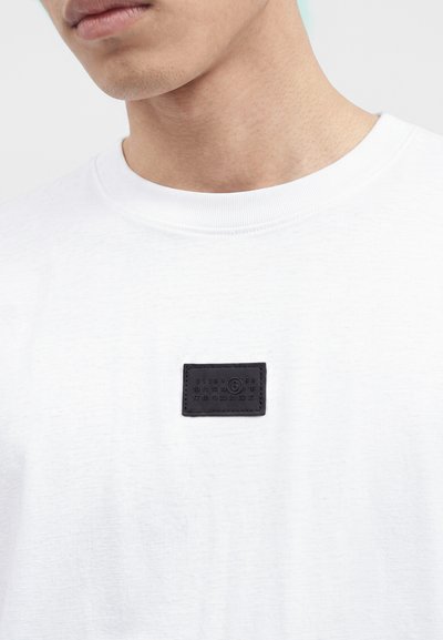 Κοντινό πλάνο ενός ατόμου που φοράει λευκό T-shirt με ένα μικρό μαύρο ορθογώνιο έμπλαστρο με αριθμούς στο στήθος.