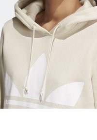 Osoba oblečená v krémovém mikině s velkým bílým logem Adidas trefoil a šňůrkami visícími dolů na přední straně.