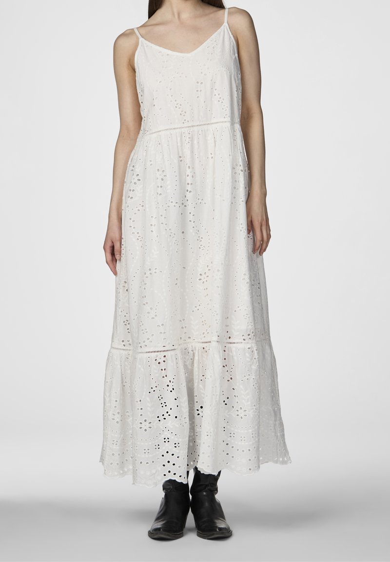 Weißes Maxikleid mit verstellbaren Spaghettiträgern, das mit einer Lochstickerei, einer taillierten Taille und einem gestuften Rock versehen ist. Kombiniert mit schwarzen Knöchelstiefeln.