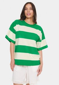 Grün- und cremegestreiftes T-Shirt aus Baumwolle mit lockerem Schnitt, kurzen Ärmeln und rundem Ausschnitt. Kombiniert mit weißen Shorts.
