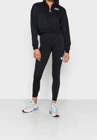 Svart kort sweatshirt med halv dragkedja och logotyp, i kombination med svarta leggings med en vit Nike-swoosh, och flerfärgad träningsskor.