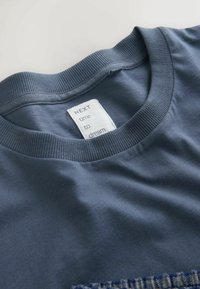 Blaules Baumwoll-T-Shirt mit geripptem Rundhalsausschnitt, ausgestattet mit einem weißen Etikett, auf dem "NEXT time to dream" steht. Glatte Textur und lässiges Design.