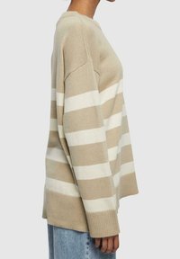 Pull en beige à rayures avec des rayures horizontales crème, poignets côtelés, large encolure et tricot texturé. Porté avec un jean bleu clair.