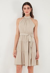 Beige halterneck mekko, joka on valmistettu kevyestä kankaasta; siinä on kerrottu kaula-aukko, vyötärönauha ja kerrostettu hame, mikä luo virtaavan siluetin.