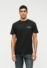 Juodas medvilninis marškinėlis su apvalia iškirpte, trumpomis rankovėmis ir mėlynu piešiniu ant krūtinės. Dėvimas su tamsiai mėlynomis džinsais.