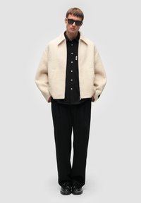 Kremowa kurtka z teksturowanej wełny z kołnierzem, kieszeniami po bokach i czarnymi guzikami, noszona na czsarną koszulę z guzikami i luźne czarne spodnie.