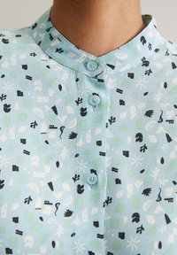 Light blue blouse featuring a collar, button closure, and a pattern of black, white, and green shapes, including leaves and geometric forms.