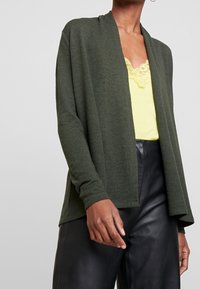 Offen vorne Cardigan in Dunkelgrün, Strickstruktur, langärmlig, kombiniert mit einem hellgelben Top, getragen über schwarzen Lederhosen.