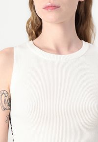 Hvit, ribbet tanktop med rund hals. Stoffet har en glatt tekstur. På siden er det en tekstdesign i svart.