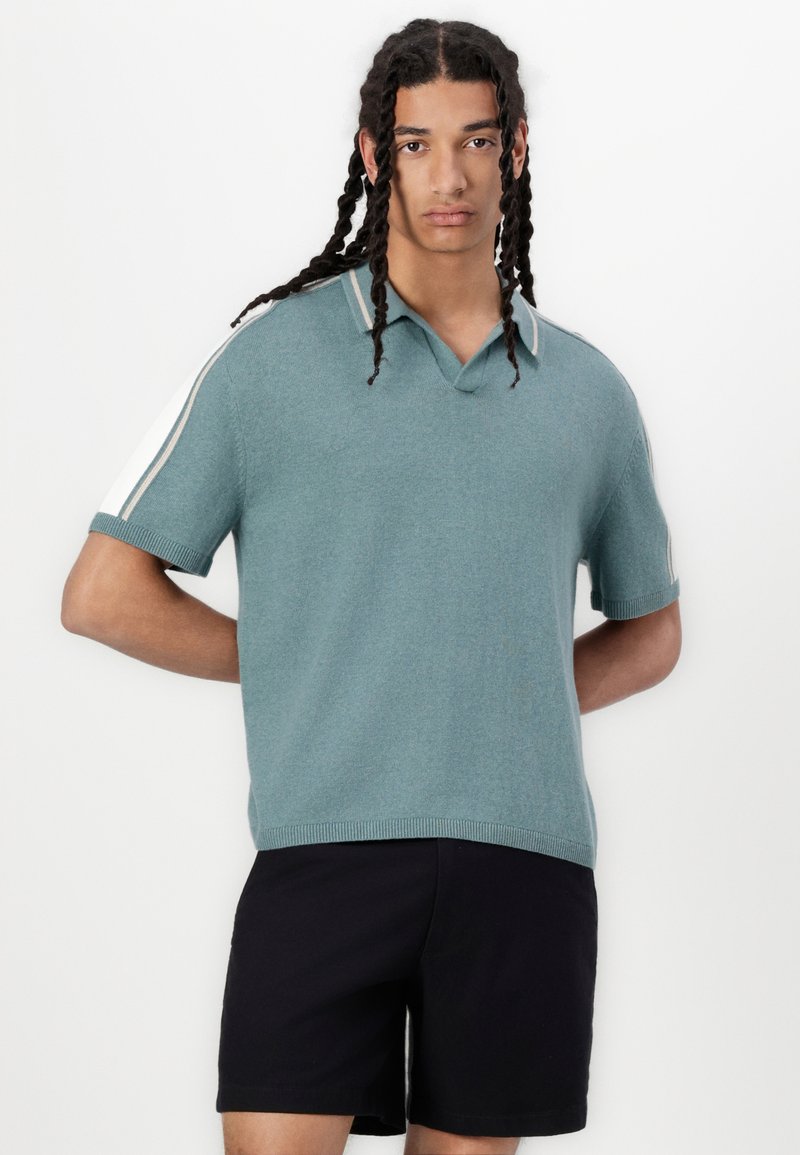 Polo en tricot à manches courtes, de couleur teal clair, avec un col en V et des accents blancs sur les épaules, associé à un short noir. Texture douce.
