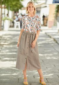 Blümchenbluse mit kurzen Ärmeln, ergänzt durch einen beigen Rock mit Knöpfen und einem Bindegürtel. Gelbe Sandalen runden das Outfit ab.