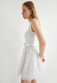 Abito bianco in pizzo con ricami floreali e vita aderente, caratterizzato da una cintura bianca liscia e una gonna a campana. Tessuto simile alla seta.