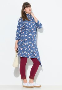 Blaue Tunika mit floralem Print, asymmetrisches Design, kombiniert mit bordeauxfarbenen Leggings mit Spitze und weißen Espadrilles, gehalten mit einer geflochtenen Tasche.