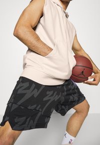 Light beige sleeveless hoodie with front pocket; black athletic shorts featuring grey zigzag patterns; holding a brown basketball.