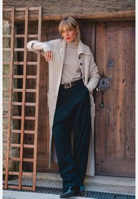 Long cardigan beige à mailles texturées et boutons, porté sur un col roulé léger. Pantalon noir à jambes larges et chaussures noires élégantes. Fond en bois.
