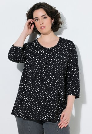 Femme aux cheveux courts bouclés foncés portant un top noir à manches longues avec des motifs triangulaires blancs et un pantalon gris sur fond blanc uni.