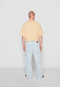 Camiseta de gran tamaño en beige claro combinada con jeans cargo azul claro que presentan bolsillos tipo parche y detalles de logotipo. Llevado con zapatillas negras y blancas.