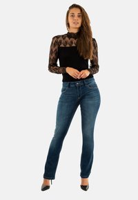 Top de encaje negro con mangas largas, cuerpo ajustado y cuello redondo, combinado con jeans de mezclilla azul de pierna recta y tacones altos negros.