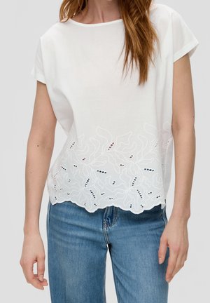 Kvinde iført en hvid kortærmet bluse med blomstret blonde ved kanten, kombineret med blå jeans, stående mod en ensfarvet baggrund.