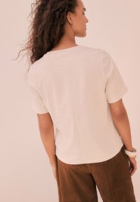 T-shirt corto a maniche corte beige chiaro con taglio dritto e colletto a girocollo, abbinato a pantaloni corti in suede marrone. Tessuto semplice e liscio, senza fantasie.