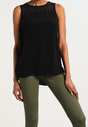 Ärmelloses schwarzes Top mit transparentem Einsatz über der oberen Brust, getragen mit eng anliegenden olivgrünen Hosen an einer stehenden Person.