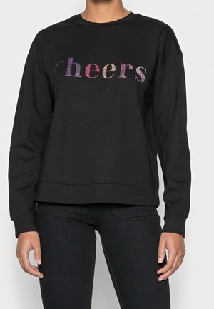 Osoba nosząca czarną bluzę z wielokolorowymi literami z kryształków tworzącymi napis „cheers” na piersi.