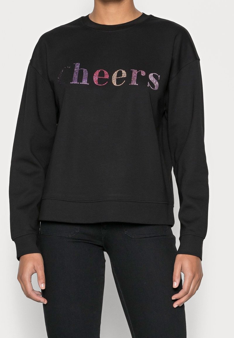 Personne portant un sweat-shirt noir avec des lettres en strass multicolores formant le mot « cheers » sur la poitrine.
