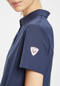 Mörknavy piké med krage, korta ärmar och Rossignol-logotyp på vänster ärm. Tyget verkar jämnt och ventilerande.