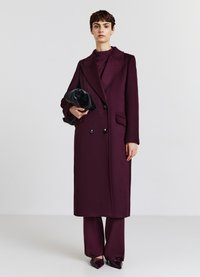 Cappotto lungo doppiopetto in lana bordeaux scuro, dotato di rever a punta, tasche laterali e abbinato a una pochette nera e pantaloni coordinati.