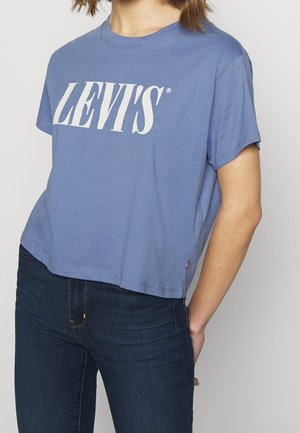 Personne portant un t-shirt court bleu Levi's et un jean bleu foncé avec une main derrière le dos, sur un fond clair uni.