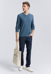 Blauwe v-hals trui met lange mouwen, gedragen met donkere spijkerbroek en witte sneakers. Houdt een beige boodschappentas met een logo vast, staand tegen een effen achtergrond.