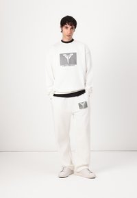 Hvid sweatshirt med grafisk tryk, parret med matchende hvide joggingbukser. Begge beklædningsgenstande har en blød tekstur og markante sorte accenter.