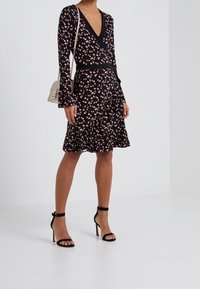 Kleid mit floralem Muster, langen Ärmeln, schwarzem Grund, rosa Blumen, ausgestelltem Saum und Rüschendetails. Kombiniert mit schwarzen Riemchen-Heels.